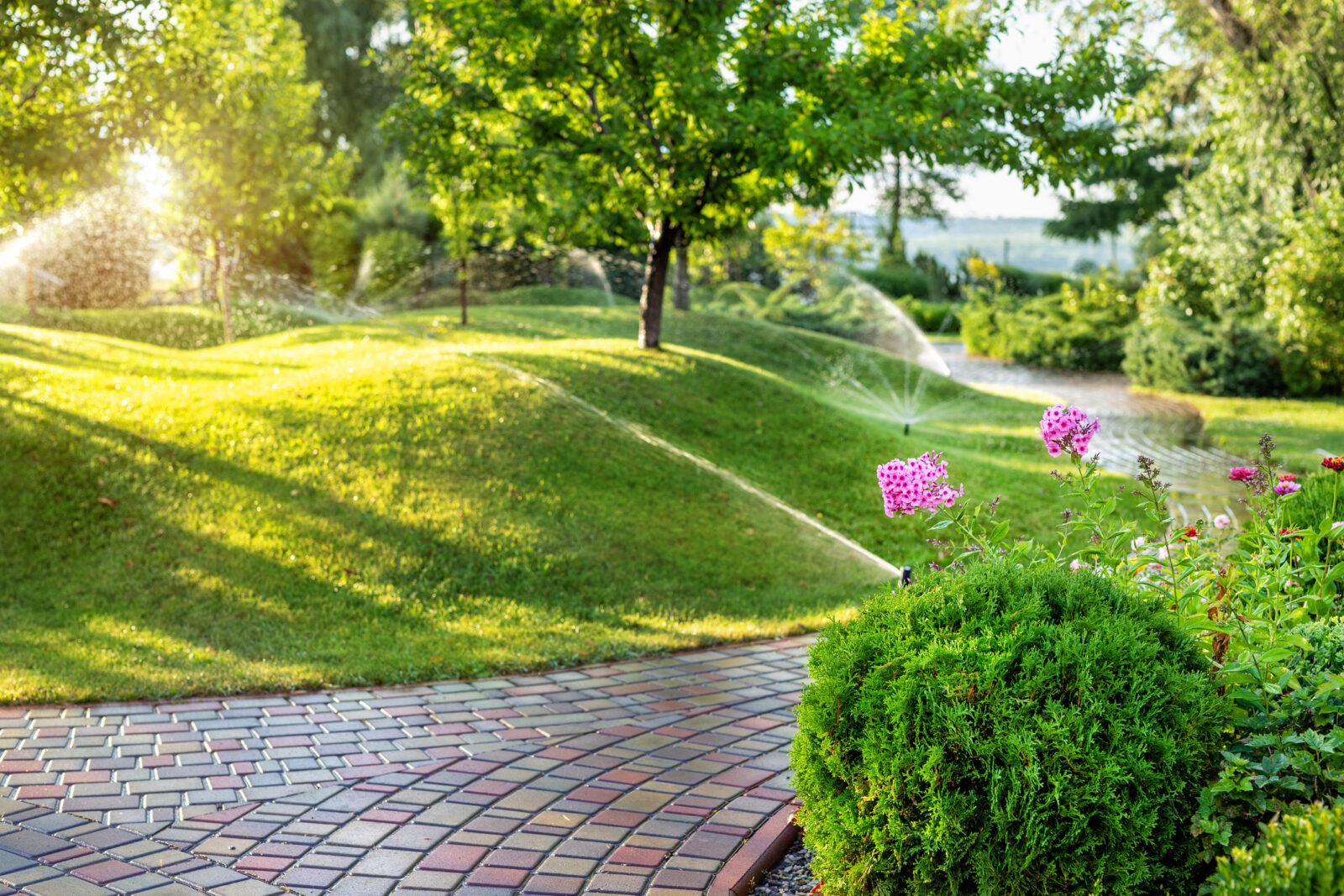 A picture of a lush green lawn and a flower garden divided by a brick walkway, to illustrate a blog post about the importance of sprucing up your sprinkler system. Sprinklers waste thousands of gallons of water a year and lead to higher water bills when they are not properly adjusted.